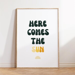 May include: A framed print with the text "HERE COMES THE SUN" in a dark green font. The word "SUN" is in a gradient of yellow and orange. A small yellow half-circle is below the text.
