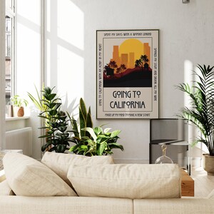 May include: A framed art print with the text "Going to California" in a retro style. The artwork features a sunset over a cityscape with palm trees. The print includes additional text around the border, and is displayed in a bright, airy room with plants and a sofa.
