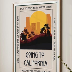 May include: A framed art print with a California theme. The artwork features a sunset over a city skyline with palm trees and the text "Going to California". The print has a beige background and a wooden frame.