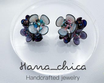 Peut inclure: Une paire de boucles d'oreilles en forme de fleurs à plusieurs couches avec des pétales translucides blancs, violets et bleu irisé. Le centre de chaque fleur présente de petites perles groupées. Le texte Hama_chica Handcrafted jewelry est visible.