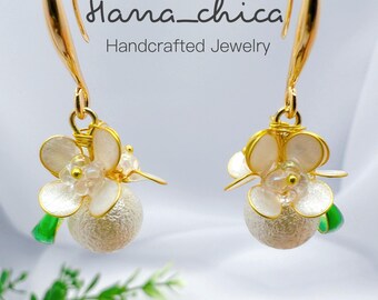 Peut inclure: Une paire de boucles d'oreilles à crochet dorées avec des breloques en forme de fleurs blanches, des perles sphériques texturées et de petits accents verts. Le texte "Hana_chica Handcrafted Jewelry" est visible au-dessus des boucles.