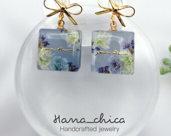 Peut inclure: Une paire de boucles d'oreilles carrées en résine contenant des fleurs séchées bleues et blanches. Chaque boucle est surmontée d'un nœud en métal doré et porte l'inscription dorée en cursive "jour blanc" sur la face avant du bloc de résine.
