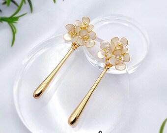 Peut inclure: Une paire de boucles d'oreilles pendantes dorées composées de grappes de petits pétales de fleurs blancs translucides avec des centres jaunes, fixées à des barres métalliques dorées allongées. Le texte Hana_chica Handcrafted jewelry est visible.