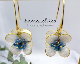 Peut inclure: Une paire de boucles d'oreilles à crochet dorées avec des breloques en forme de fleur, des pétales blancs nacrés et un centre composé de petites perles bleues. Le texte Hana_chica Handcrafted jewelry est visible au-dessus.