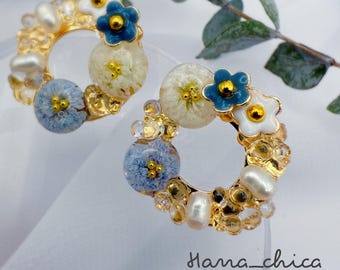Peut inclure: Une paire de boucles d'oreilles circulaires avec des montures en métal doré ornées d'accents floraux blancs et bleus, de petites perles et de perles transparentes. Le texte Hana_chica Handcrafted jewelry est visible en bas à droite.
