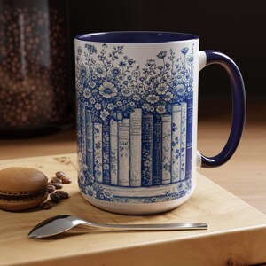 May include: White ceramic mug with a blue floral and book design. The mug features a blue handle and rim. A silver spoon and a macaron are on a wooden surface next to the mug.