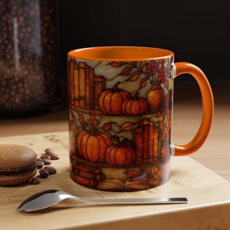May include: Orange ceramic mug with a fall-themed design featuring pumpkins, books, and flowers. The interior and handle are solid orange. The mug sits on a wooden surface with a spoon and a pastry.