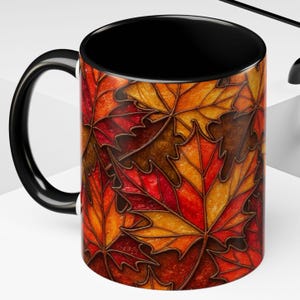 May include: A ceramic mug with a black interior and handle. The exterior features a repeating pattern of autumn maple leaves in shades of red, orange, and brown. The mug is designed for hot beverages.