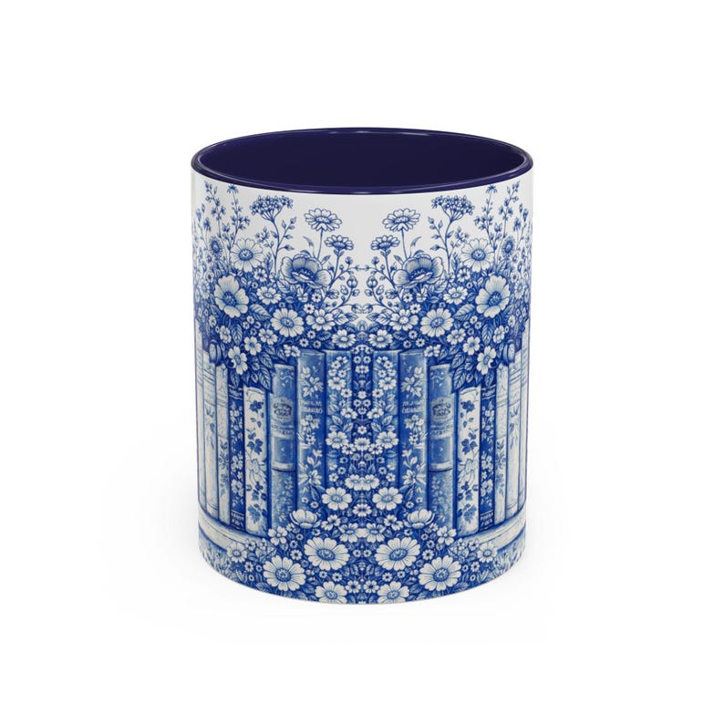 May include: White ceramic mug with a blue floral and book design. The mug features a dark blue interior and a wraparound pattern of white flowers, books, and foliage in a blue and white color scheme. The mug is cylindrical.