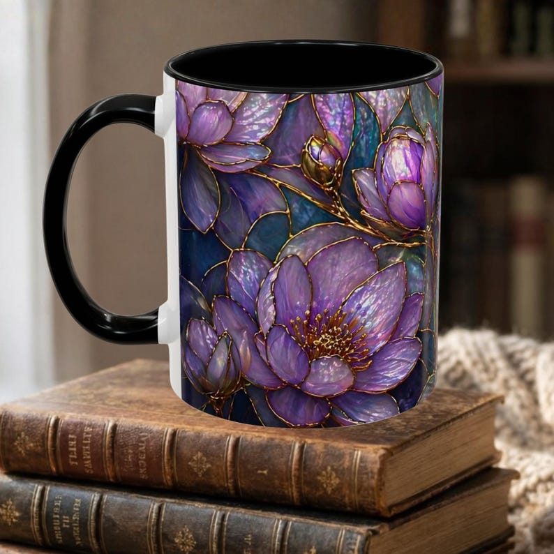 May include: A ceramic coffee mug with a black handle and interior. The exterior features a floral design with purple flowers, gold outlines, and blue-green leaves. The mug is sitting on a stack of old books.