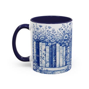 May include: White ceramic mug with a navy blue interior and handle. The exterior features a blue and white floral and book design. The design wraps around the mug, creating a cohesive aesthetic. The mug is suitable for hot or cold beverages.