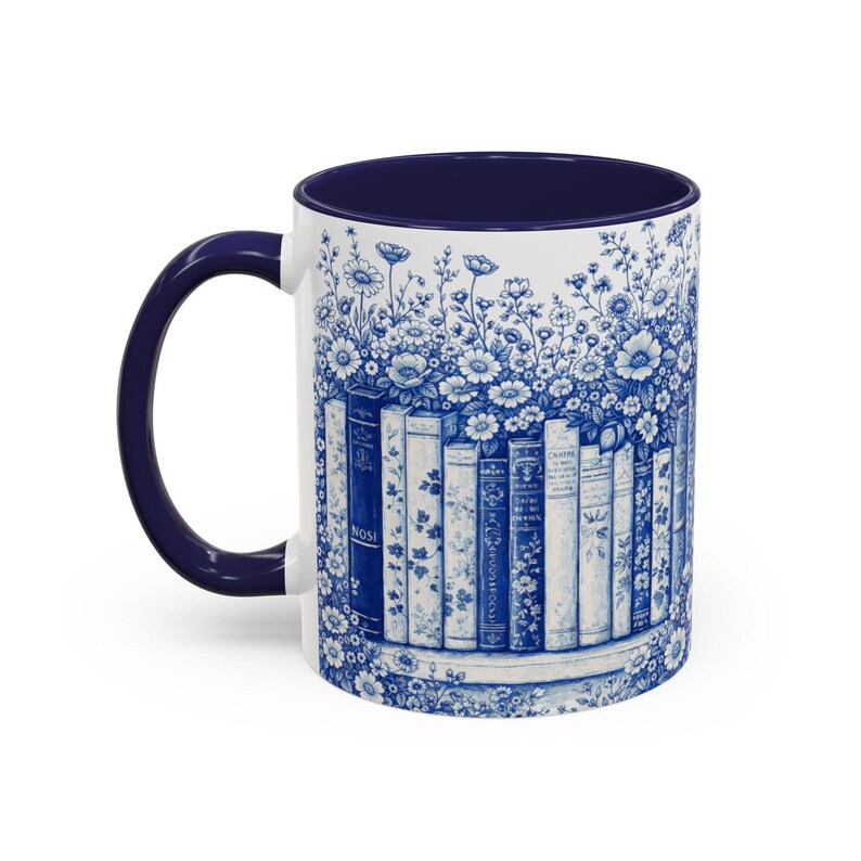 May include: White ceramic mug with a navy blue interior and handle. The exterior features a blue and white floral and book design. The design wraps around the mug, creating a cohesive aesthetic. The mug is suitable for hot or cold beverages.