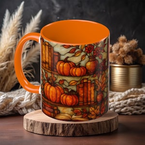 May include: Orange ceramic mug with a fall-themed design. The mug features a stained-glass style illustration of pumpkins, books, and autumn leaves. The interior and handle of the mug are solid orange.