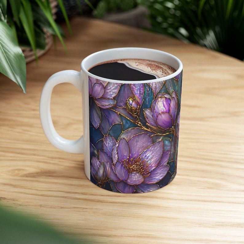 May include: White ceramic coffee mug featuring a floral design with purple and gold accents. The flowers are set against a dark blue background. The mug is filled with dark coffee and has a white handle.
