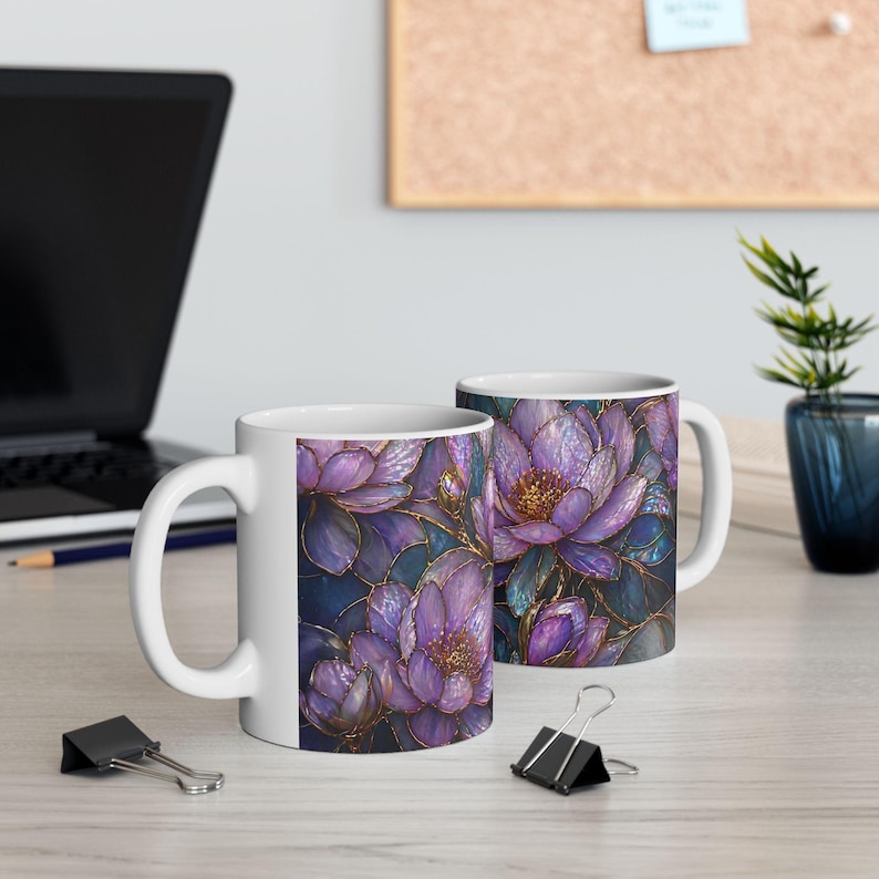 May include: Two white ceramic mugs with a floral design. The design features purple flowers with gold accents against a dark blue background. The mugs are on a light-colored wooden surface, with a laptop and office supplies in the background.