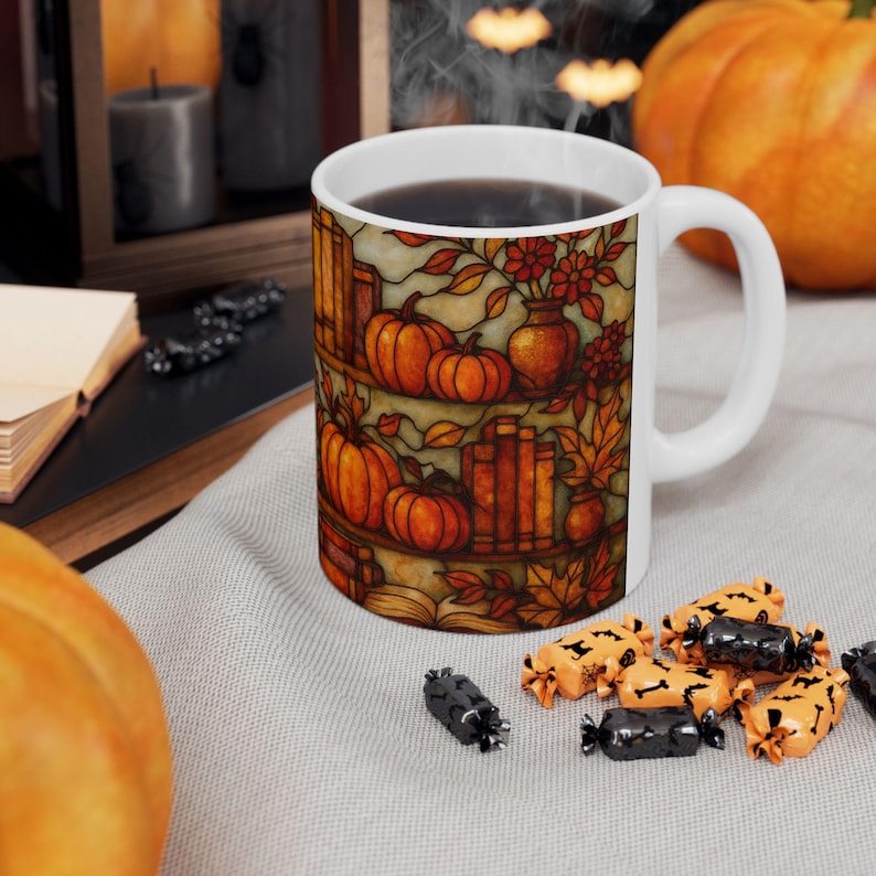 May include: White ceramic mug with a colorful stained-glass design featuring pumpkins, books, and fall foliage. The mug is filled with dark liquid and has a white handle. Candy corn and black candies are scattered around the mug.