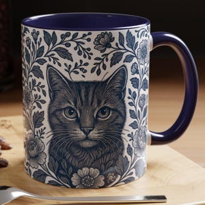 May include: A ceramic mug with a blue interior and handle. The exterior features a detailed illustration of a cat's face surrounded by floral and leaf patterns in shades of blue and white. The mug is on a wooden surface.