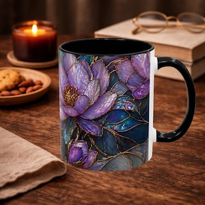 May include: A ceramic coffee mug with a black interior and handle, featuring a floral design in shades of purple, blue, and gold. The flowers have a stained glass effect. The mug is white on the exterior and sits on a wooden surface.