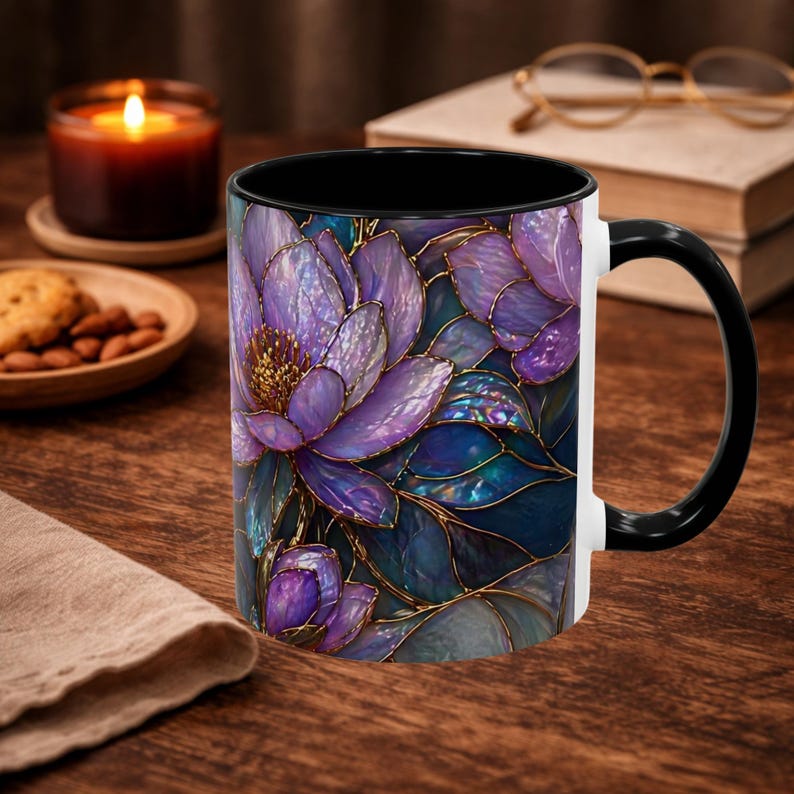 May include: A ceramic coffee mug with a black interior and handle, featuring a floral design in shades of purple, blue, and gold. The flowers have a stained glass effect. The mug is white on the exterior and sits on a wooden surface.