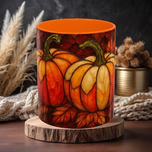 May include: A cylindrical ceramic mug with an orange interior and a stained-glass pumpkin design. The pumpkins are orange and yellow with green stems, set against a dark red background. The mug sits on a wooden coaster.