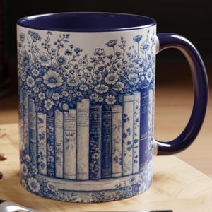 May include: A ceramic mug with a blue interior and handle, decorated with a white and blue floral and book design. The mug's exterior displays a detailed illustration of flowers and a row of vintage books.