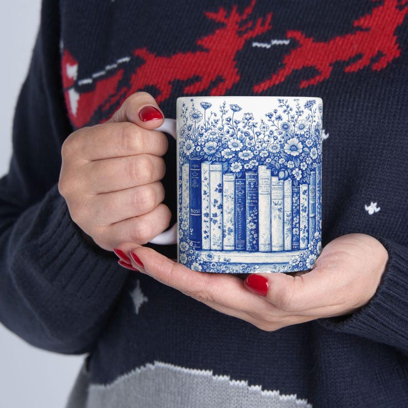May include: White ceramic mug with a blue and white floral and book design. The mug features a row of books with blue spines and floral accents. The handle is white.