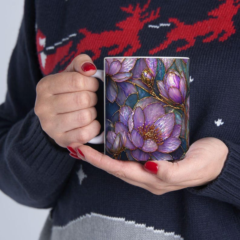 May include: White ceramic mug featuring a floral design with purple and gold accents. The flowers are detailed with a stained-glass effect, set against a dark blue background. The mug is held by a person wearing a navy blue sweater.