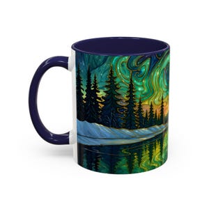 May include: A white ceramic coffee mug with a dark blue handle and interior. The mug features a vibrant illustration of a forest scene with a lake reflecting the aurora borealis in shades of green, gold, and blue.