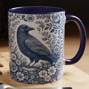 May include: A ceramic mug with a dark blue interior and handle. The exterior features a detailed illustration of a raven surrounded by floral patterns in shades of blue and white. The mug is designed for hot beverages.