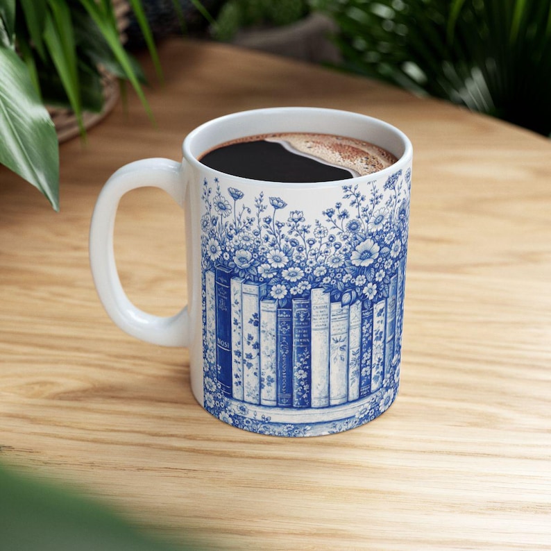 May include: White ceramic coffee mug with a blue floral and bookshelf design. The mug is filled with dark coffee. The design features blue flowers and a row of books. The mug has a white handle and is sitting on a wooden surface.