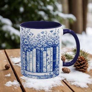 May include: White ceramic mug with a blue floral and book design. The mug features a navy blue interior and handle. The design includes a row of books with floral accents, creating a vintage aesthetic. The mug is set on a wooden surface with snow.