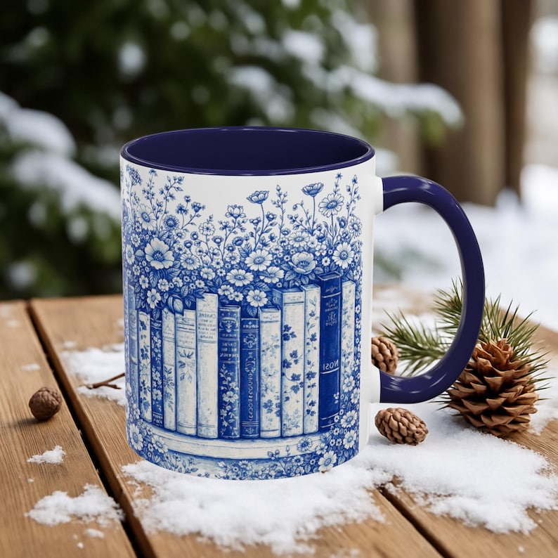 May include: White ceramic mug with a blue floral and book design. The mug features a navy blue interior and handle. The design includes a row of books with floral accents, creating a vintage aesthetic. The mug is set on a wooden surface with snow.