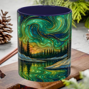 May include: A ceramic mug with a vibrant landscape design. The artwork features a green and blue aurora borealis over a forest and lake scene. The mug has a dark blue interior and a gold outline on the design.