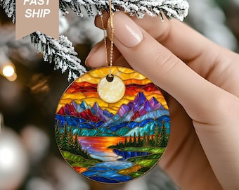 May include: Round Christmas ornament with a stained-glass design featuring a mountain landscape with a lake and sunset. The ornament has a gold hanging loop and is held by a person. The text "FAST SHIP" is visible.