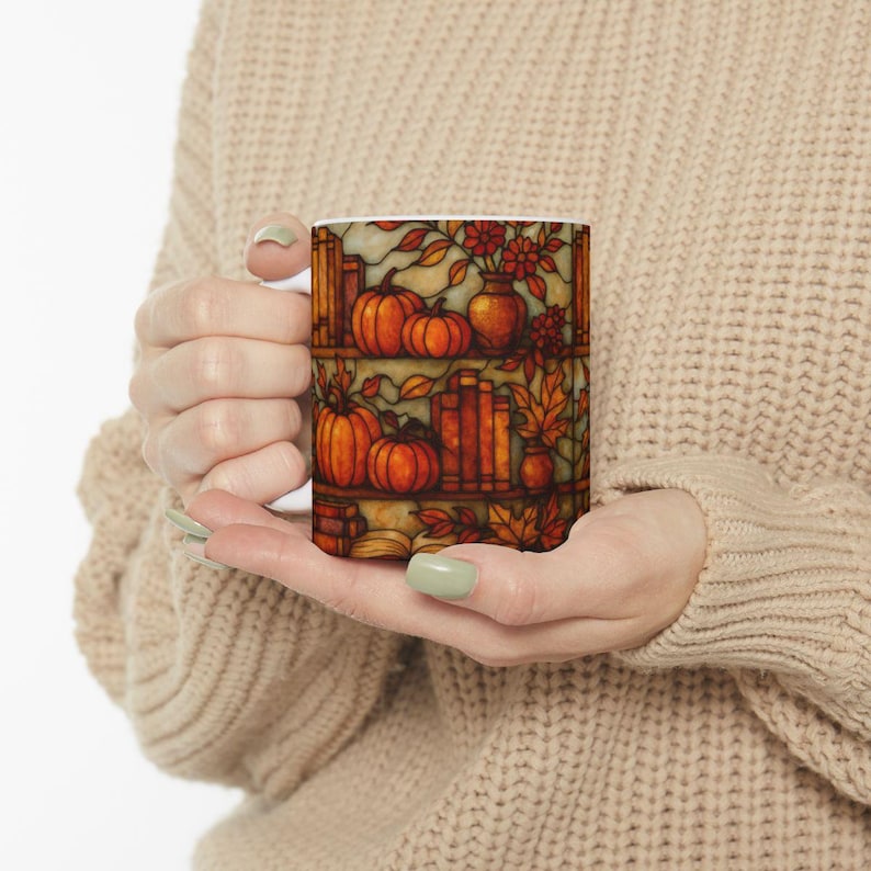 May include: White ceramic mug featuring a fall-themed design. The design includes pumpkins, books, vases of flowers, and autumn leaves. The artwork is in warm orange, brown, and yellow tones, evoking a cozy, seasonal feel. The mug is held by a person.