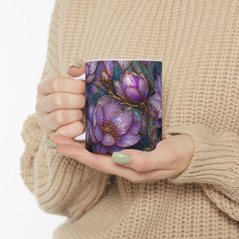 May include: White ceramic mug featuring a detailed floral design. The design showcases purple flowers with gold accents against a dark blue background. The mug is held by a person wearing a beige sweater.