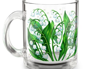 May include: Clear glass mug featuring a detailed illustration of lily of the valley flowers. The design includes white bell-shaped flowers, green leaves, and small pearl-like accents. The mug has a clear handle and base.