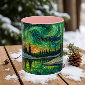 May include: A ceramic mug with a pink interior and a wrap-around design featuring a landscape with a forest, lake, and swirling green and gold aurora borealis. The mug is set on a wooden surface with snow.