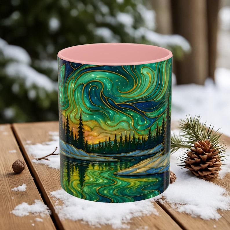 May include: A ceramic mug with a pink interior and a wrap-around design featuring a landscape with a forest, lake, and swirling green and gold aurora borealis. The mug is set on a wooden surface with snow.