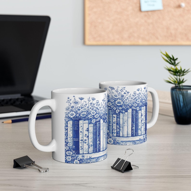 May include: Two white ceramic mugs with a blue floral and bookshelf design. The mugs feature a wraparound pattern of blue flowers and a row of books. The mugs are on a light-colored surface.