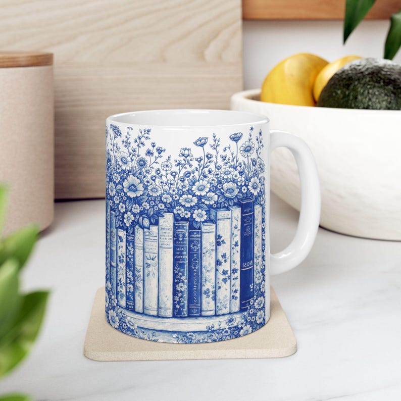 May include: White ceramic mug with a blue floral and book design. The mug features a row of books with floral accents and a handle. The design wraps around the mug, creating a cohesive look.