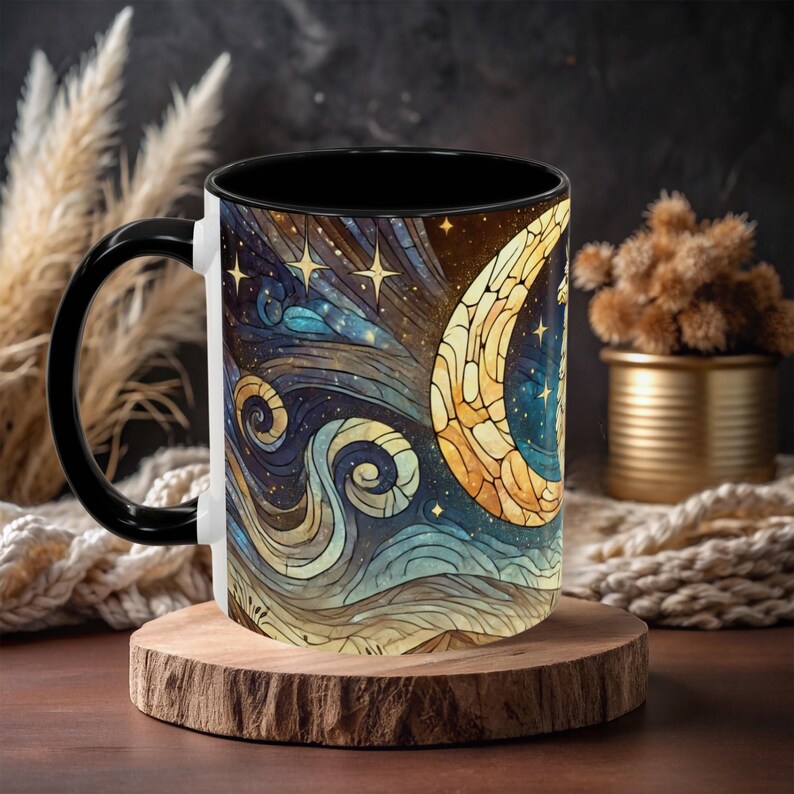 May include: A white ceramic mug with a black handle and a black rim. The mug features a colorful stained glass design of a crescent moon and stars against a dark blue background.