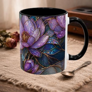 May include: A ceramic coffee mug with a floral design. The mug features a black interior and handle, with a white exterior. The floral design is a mosaic of purple and blue flowers with gold outlines. A spoon rests on the table.