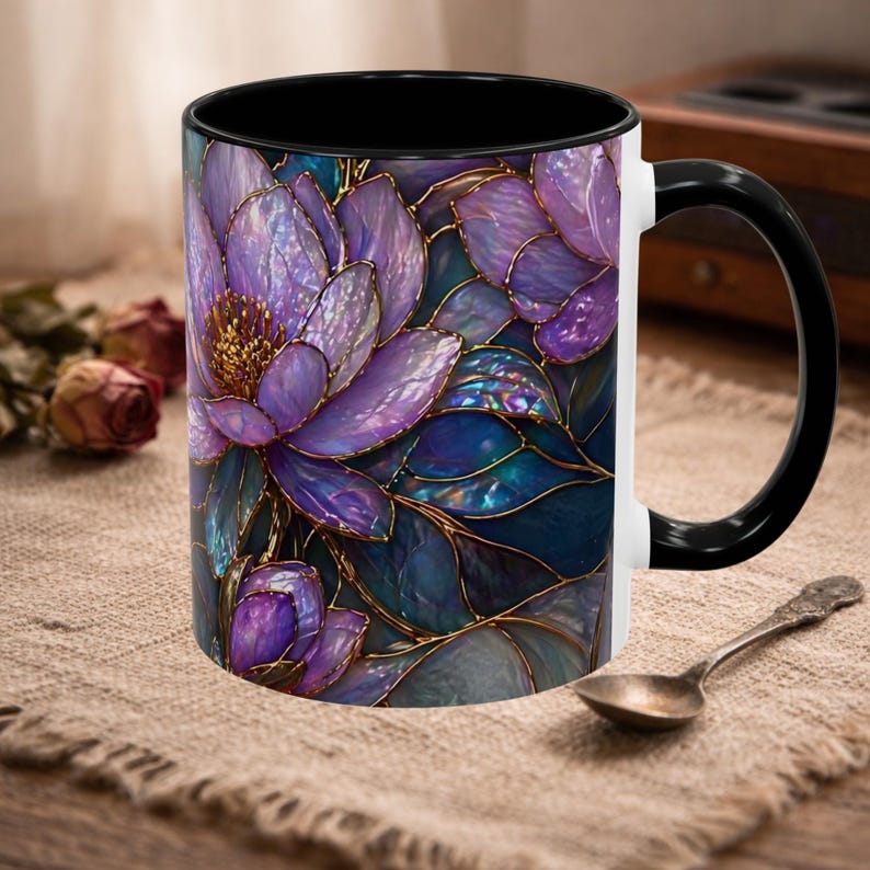 May include: A ceramic coffee mug with a floral design. The mug features a black interior and handle, with a white exterior. The floral design is a mosaic of purple and blue flowers with gold outlines. A spoon rests on the table.