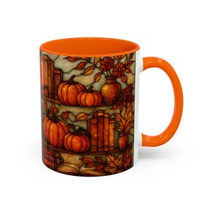 May include: White ceramic mug with an orange interior and handle. The mug features a stained-glass style design of pumpkins, books, and fall foliage. The design includes orange, brown, and yellow colors.