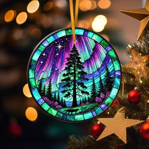 May include: Round Christmas ornament with a stained-glass design. The ornament features a vibrant scene of a starry night sky with the aurora borealis, a tall tree, and a forest. The ornament is hanging from a gold ribbon.