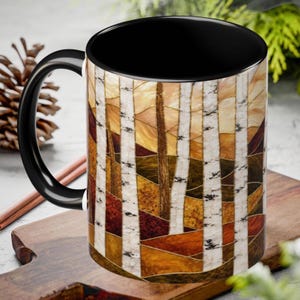 May include: A ceramic mug with a black handle and interior, featuring a wrap-around stained glass style print of a forest with white birch trees against a background of warm autumn colors including shades of orange, yellow, brown, and deep red.