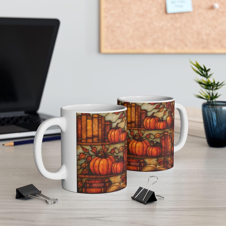 May include: Two white ceramic mugs with a fall-themed design. The design features pumpkins, books, and autumn leaves in shades of orange, brown, and yellow. The mugs are on a light-colored surface with a laptop and office supplies in the background.