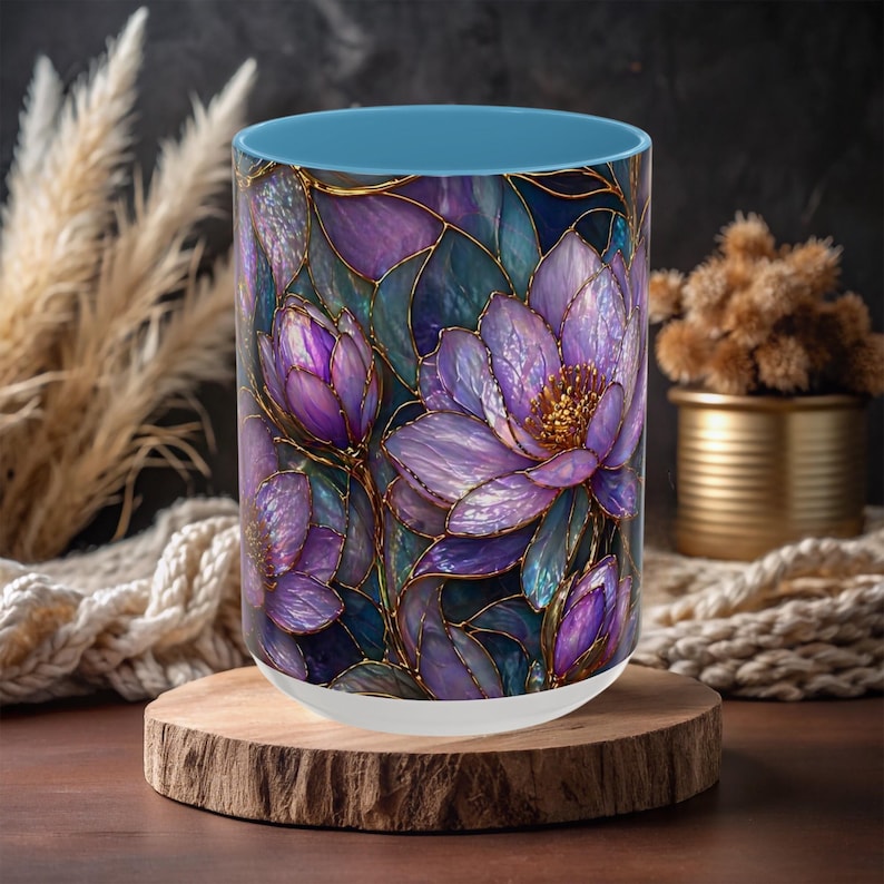 May include: A cylindrical glass mug with a blue rim and a white base. The mug features a stained-glass style floral design in shades of purple, blue, and gold. The mug is displayed on a round wooden coaster.