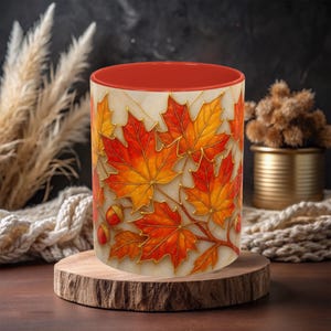 May include: A ceramic mug with a red interior and a cream-colored exterior decorated with vibrant autumn leaves in shades of red, orange, and yellow. The leaves are accented with gold-colored outlines and accompanied by acorns. The mug is displayed on a wooden slice.